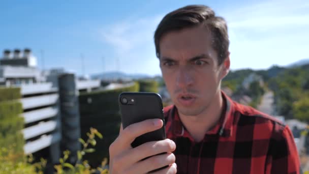 Az ember dühös az üzenetre, amit az okostelefonon olvasott. Agresszív fiatalember kockás ingben Okostelefon használata szabadban városi háttér. — Stock videók