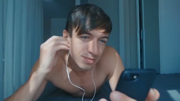 一个英俊的裸体男人，带着耳机躺在床上，在智能手机上听音乐 — 图库视频影像