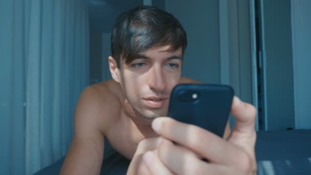 Handsome naked smiling man with headphones lying in bed at home and scrolling on smartphone — Stok video