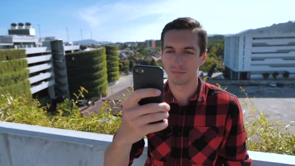 Hombre atractivo en camisa a cuadros usando teléfono inteligente que toca la pantalla al aire libre en el fondo urbano de la ciudad. Dispositivos, personas y concepto juvenil moderno — Vídeos de Stock