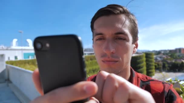 Hombre joven desplazándose por la pantalla del teléfono inteligente. Empresario usando su teléfono inteligente y pantalla móvil afuera en el techo en el fondo urbano de la ciudad — Vídeos de Stock