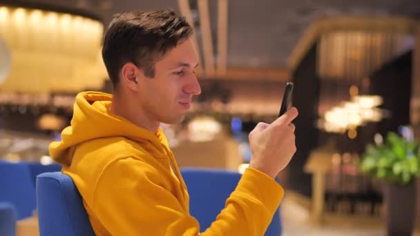 Jovem homem feliz com capuz amarelo usando smartphone no café acolhedor à noite. Comunicação, negócios, viagens, namoro conceito . — Vídeo de Stock