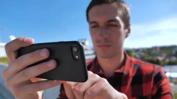 スマートフォンでビデオを見ている男。都市部の屋根の上に彼のスマートフォンでメディアを見て若い男のクローズアップ. — ストック動画