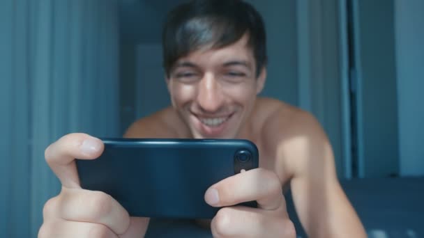 Yakışıklı, gülümseyen adam evde yatağında uzanmış akıllı telefondan mesajlar okuyor. Genç bir erkek sabah evde, yatak odasında cep telefonu kullanıyor.. — Stok video