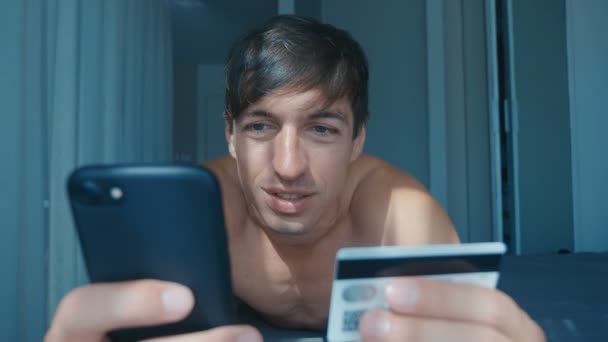 침대에 누워 있는 집에서 스마트폰 과 신용 카드를 사용하여 온라인으로 구매하는 벌거벗은 남자. 신용 카드를 들고 온라인으로 구매하고 스마트폰을 사용하는 행복 한 남성. — 비디오
