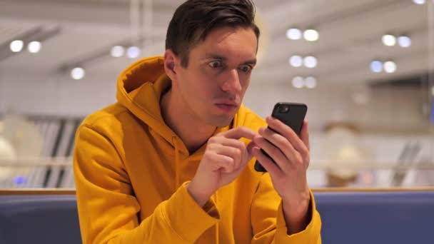 Jongeman met een gele hoodie zit in een winkelcentrum en maakt gebruik van een moderne smartphone — Stockvideo