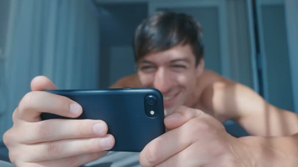 Feliz hombre sonriente viendo vídeo en el teléfono inteligente acostado en la cama en casa. Masculino riendo desnudo usando el juego de teléfono inteligente — Vídeos de Stock