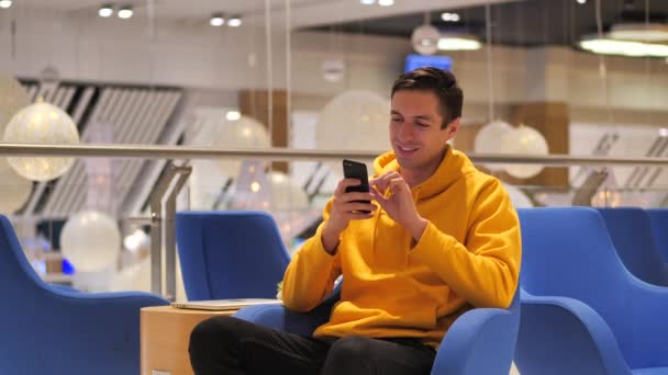 Junger lächelnder glücklicher Mann in gelbem Kapuzenpulli mit Smartphone in gemütlichem Café zur Abendzeit. — Stockvideo