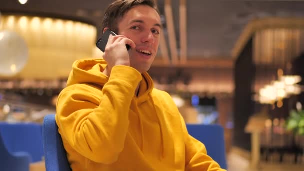 Sonriente joven hombre milenario hablando en el teléfono móvil en acogedora cafetería grande . — Vídeos de Stock