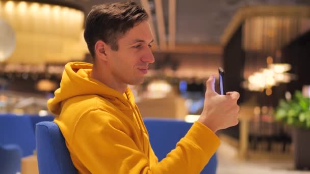 Молодой человек в желтой толстовке сидит в торговом центре кафе и пользуется современным смартфоном — стоковое видео