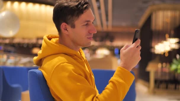 Joven con capucha amarilla se sienta en un centro comercial cafetería y utiliza un teléfono inteligente moderno — Vídeos de Stock