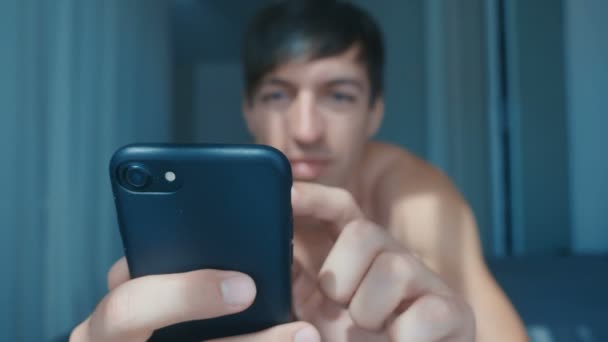 Jóképű meztelen mosolygós férfi fekszik az ágyban otthon, és az okostelefon — Stock videók
