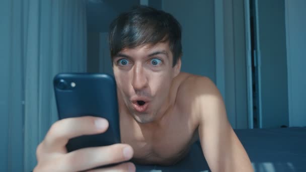 Молодий голий чоловік святкує успіх на смартфоні в ліжку вдома вранці. Емоційна реакція на перемогу . — стокове відео