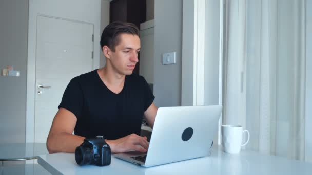 Jeune photographe utilisant un ordinateur portable traitant des images sur son ordinateur portable et buvant du café à la maison. Caméra, Disque dur externe couché à côté . — Video