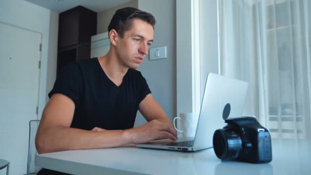 Junger Fotograf, der mit seinem Laptop Bilder bearbeitet und zu Hause Kaffee trinkt. Kamera, externe Festplatte liegen neben. — Stockvideo