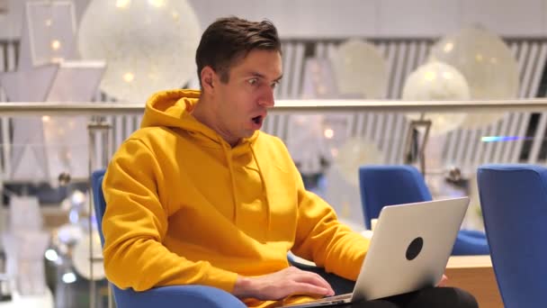 Mladý muž přemýšlí v šoku na notebooku, zatímco sedí v kavárně. Muž ve žluté mikině šokován tím, co viděl na monitoru počítače 4k 60fps — Stock video