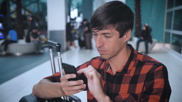 Young Man toerist in een geruit shirt met bagage op de luchthaven terminal maakt gebruik van een smartphone en wachten op boarding — Stockvideo