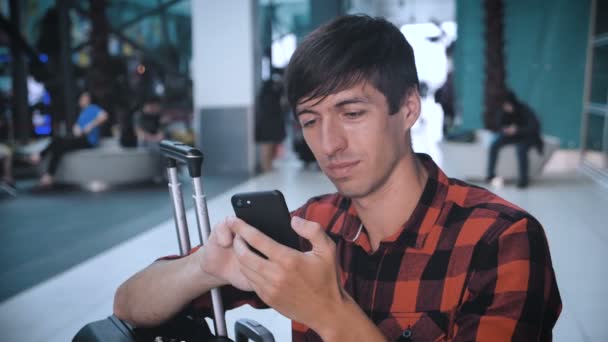 空港ターミナルで荷物のある有料シャツの男の観光客はスマートフォンを使用して搭乗を待っています。 — ストック動画