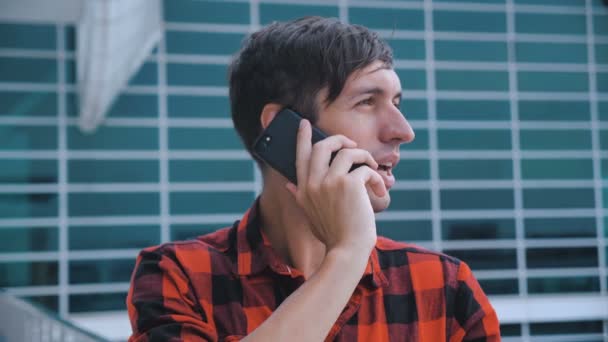 Junger attraktiver Mann, der in der Nähe eines modernen Bürogebäudes steht. Gespräche auf seinem Smartphone. männlich, der einen Anruf entgegennimmt. rotes kariertes Hemd. — Stockvideo