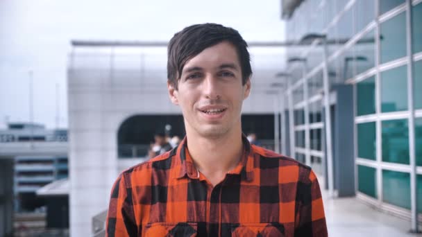 Портрет молодого человека в клетчатой рубашке, улыбающегося, смотрящего в камеру на открытом воздухе на фоне большого современного здания — стоковое видео