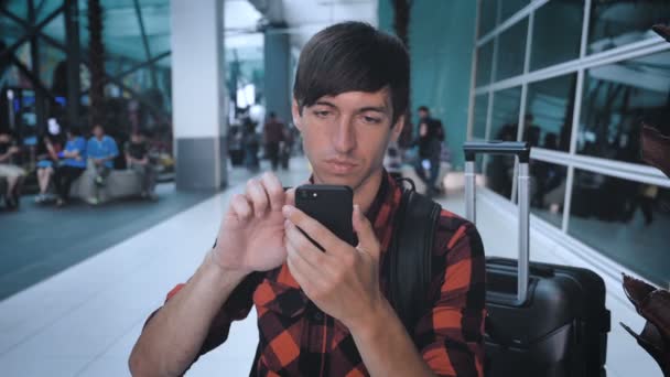 Человек турист в клетчатой рубашке с багажом на терминале аэропорта пользуется смартфоном и ждет посадки — стоковое видео