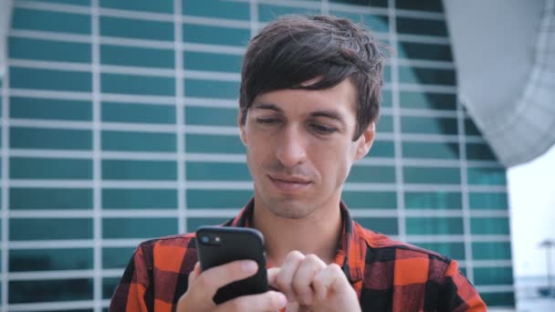 Homem atraente em pé perto de Big Modern Office Glass Building e usando Smartphone. Camisa xadrez vermelho vestido . — Vídeo de Stock