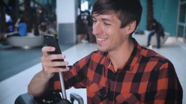Smiling Man tourist in a plaid shirt with luggage at the airport terminal uses a smartphone and waiting for boarding