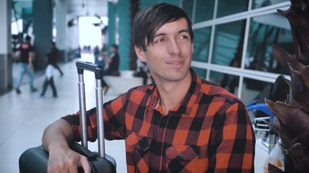 Portrait of smiling man tourist with bag luggage at the airport while waiting for boarding. Caucasian hipster male in plaid shirt in terminal of airport waiting for plane — Stock Video