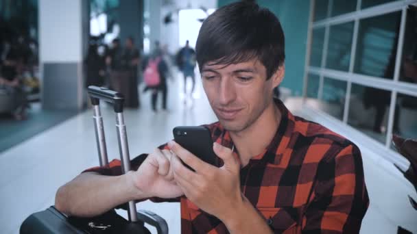 Turista di sesso maschile in camicia a quadri con bagagli al terminal dell'aeroporto utilizza uno smartphone e in attesa di imbarco — Video Stock