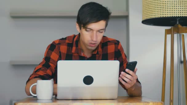 Confident busy businessman freelancer works at a laptop, drinks coffee and uses smartphone while sitting at table at home — 图库视频影像