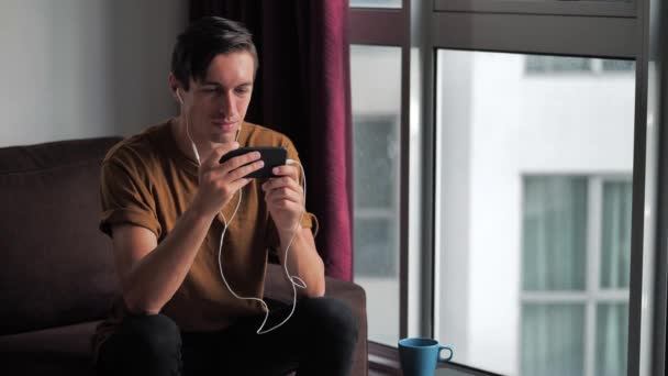 Joven con auriculares disfruta de un smartphone viendo los medios de comunicación en las redes sociales mientras está sentado en casa en el sofá — Vídeo de stock