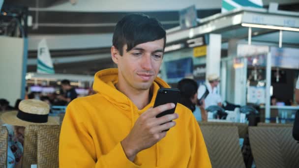 Красивый белый мужчина в желтой толстовке сидит в салоне вылета в аэропорту, ждет полета, печатает сообщение на смартфоне и улыбается — стоковое видео