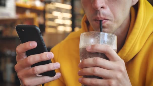 Primer plano del hombre en sudadera con capucha amarilla utilizando la pantalla táctil del teléfono inteligente en la cafetería moderna. y Beber batido o batido — Vídeos de Stock