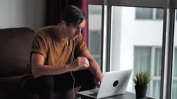 Щасливий молодий чоловік в навушниках використовує портативний комп'ютер для електронної комерції робить онлайн-покупки дивитися відеофільм, сидячи на дивані вдома — стокове відео