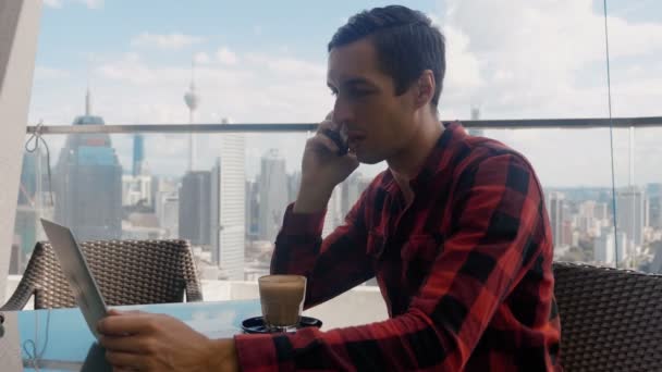 Confident business man talking on the phone and working on a laptop in a cafe on the roof of a skyscraper against the background of a big city metropolis skyscrapers. — Stock video