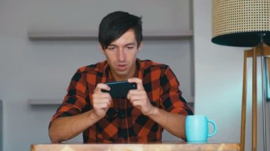 Young man using mobile phone playing video games at home. Male shows different emotions during the game.