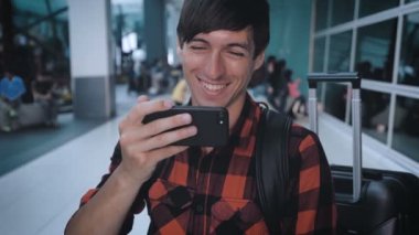 Havaalanı terminalinde ekose gömlekli gülümseyen bir turist akıllı telefondan video izleyip uçağa binmeyi bekliyor.