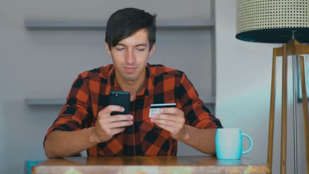 Man with credit card and phone. Young male sitting at desk at home and makes online payments through the Internet from bank card on smartphone — Wideo stockowe