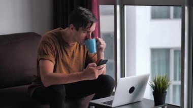 Busy man using smartphone and laptop computer for ecommerce doing online shopping watching video movie sitting on sofa at home