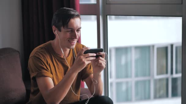 Радостный человек в наушниках наслаждается смартфоном, смотрящим медиа в социальных сетях, сидя дома на диване — стоковое видео