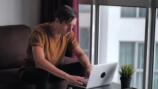 Joven guapo utiliza una computadora portátil mientras está sentado en casa en un sofá delante de una ventana . — Vídeos de Stock