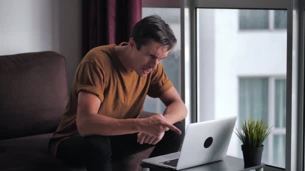 Rozzlobený problémový stresovaný muž, který má problém při používání notebooku, zatímco sedí na pohovce doma proti oknu. Podnikatel křičí, zatímco reaguje na ztrátu. Koncept nervového zhroucení. — Stock video