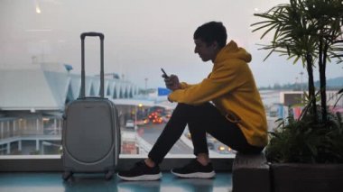 Havaalanı terminalinde bekleyen bavullu gülümseyen adam temiz pencere, gezgin akıllı telefondan mesaj atıyor ve uçağa binmeyi bekliyor.