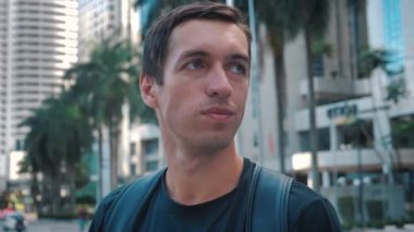 Portrait of young handsome tourist man with backpack standing outdoors alone smiling looking at camera at urban ciry background