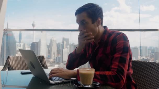 Man is unpleasantly surprised and shocked by the bad news seen on the laptop screen. Man suffers in stress and despair, while sitting at cafe on background of Big City Skyscrapers. — Wideo stockowe