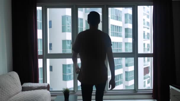 Silhouette of young man at home goes to the window of the room and looks around. The concept of dreams and success. — Stockvideo