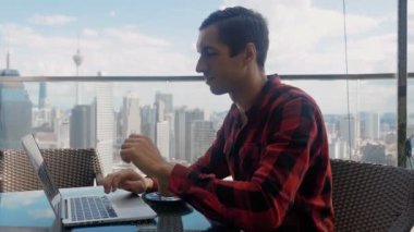 Confident business man working on a laptop and drinking coffee in cafe on the roof of a skyscraper on background of a big city metropolis. Remote work, freelance concept