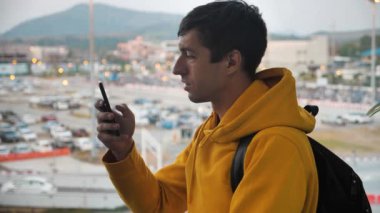 Portrait of man tourist with a backpack stands indoors in front of a large window and uses a smartphone. Travel concept.