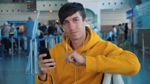 黄色のパーカーの若い男性ヒップスター空港で飛行機を待っているとヘッドフォンで彼のスマートフォンを使用して — ストック動画
