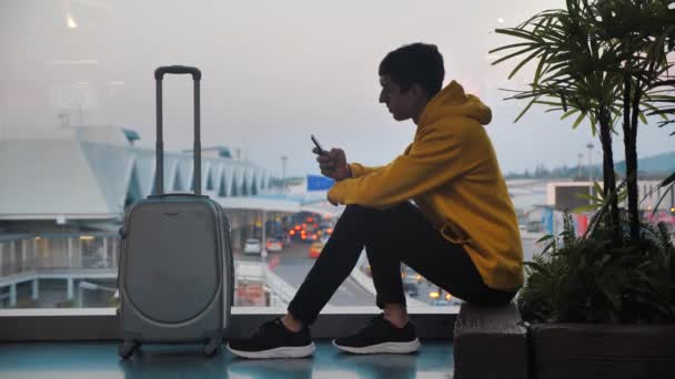 Siluet turis muda dengan bagasi menunggu di terminal bandara duduk di dekat jendela, pelancong menggunakan smartphone dan menunggu untuk boarding. Pesawat di latar belakang . — Stok Video